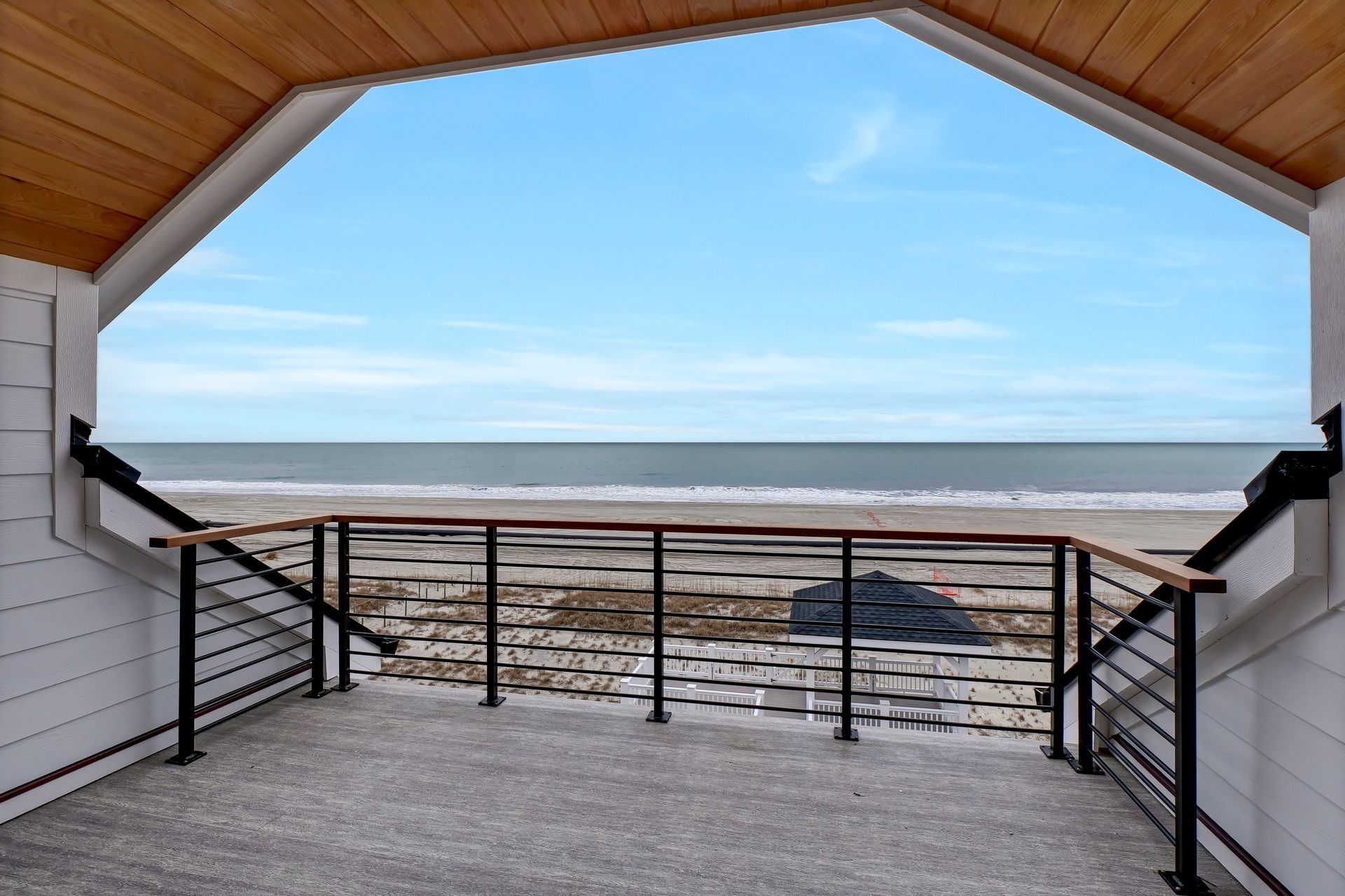 View of a beach and ocean through a modern porch with a gray floor and black railings. Blue sky.