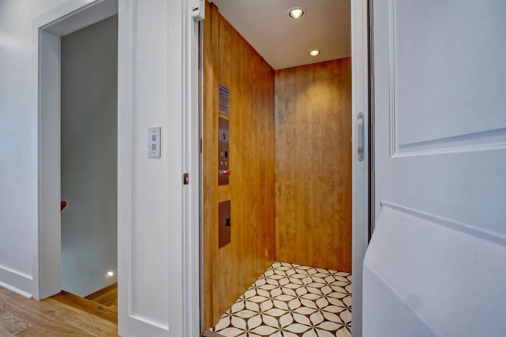 Elevator with wood paneling and patterned floor. Open door reveals controls and a stairwell.