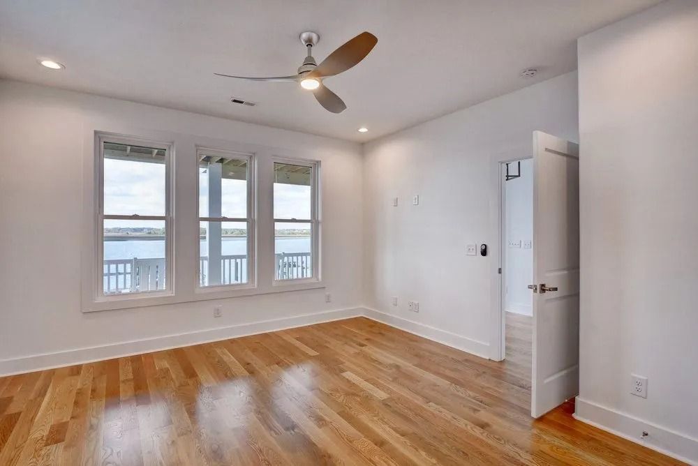 Empty bedroom with hardwood floors, three windows overlooking water, open door to another room.