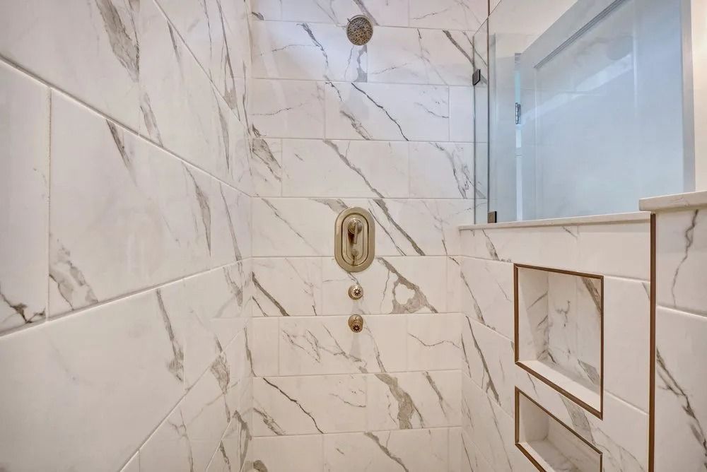 Marble-tiled shower with a rainfall showerhead, control panel, and built-in shelves. A door is to the right.