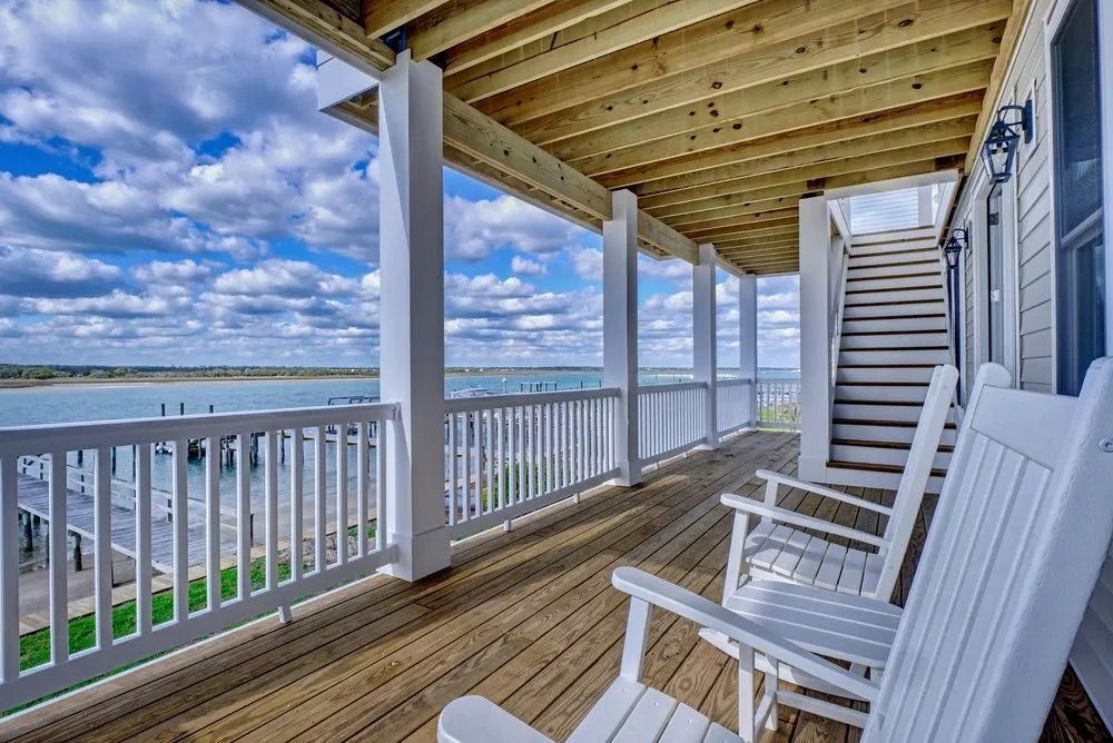 Wooden deck with white railing and rocking chairs, overlooking a waterfront on a sunny day.
