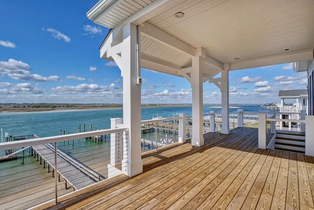 Wooden deck overlooking water, pier, blue sky with clouds. White pillars and railings on the deck.