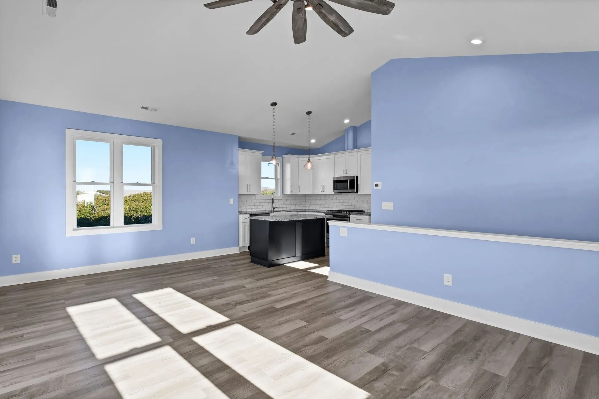 Empty living space with light blue walls, kitchen, and wood floor. Bright sunlight through the window.
