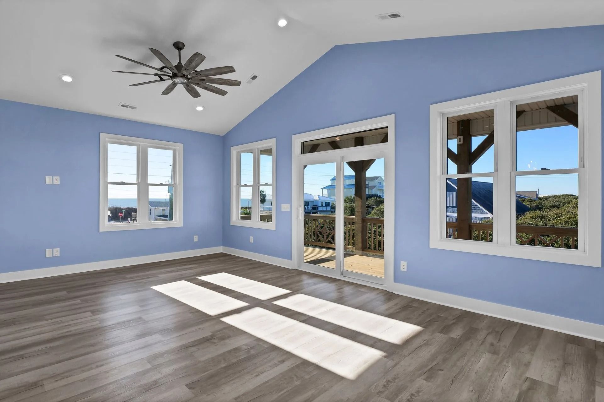 Spacious room with blue walls, gray floors, and windows overlooking a deck and ocean view; sunlight streams through.