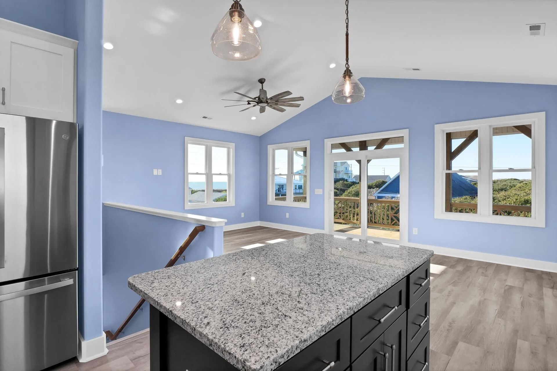 Kitchen with light blue walls, gray granite island, stainless steel refrigerator, and windows overlooking a view.