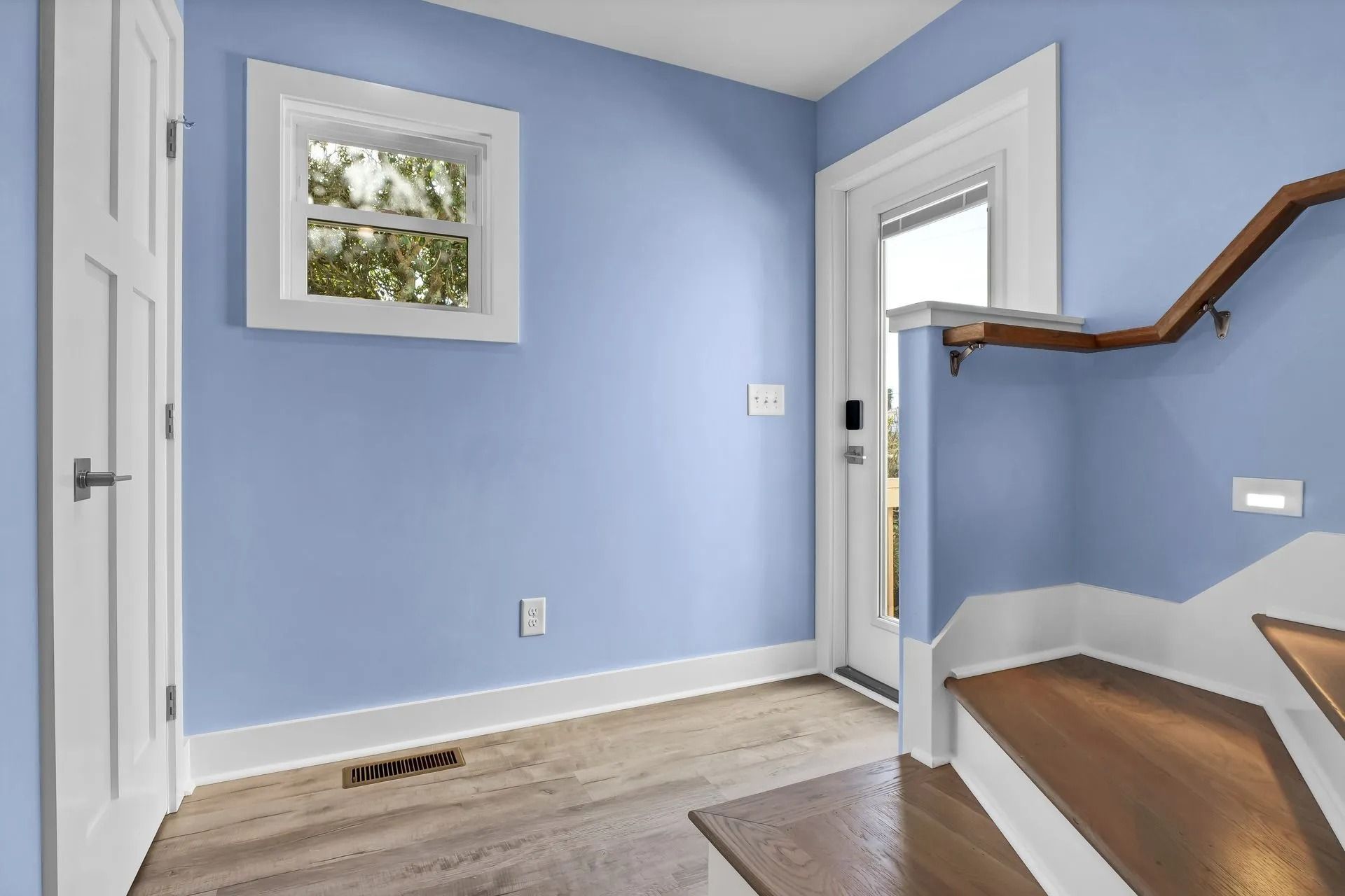 Blue-walled entryway with white trim, door, and window. Wooden stairs with a brown handrail and light colored floor.