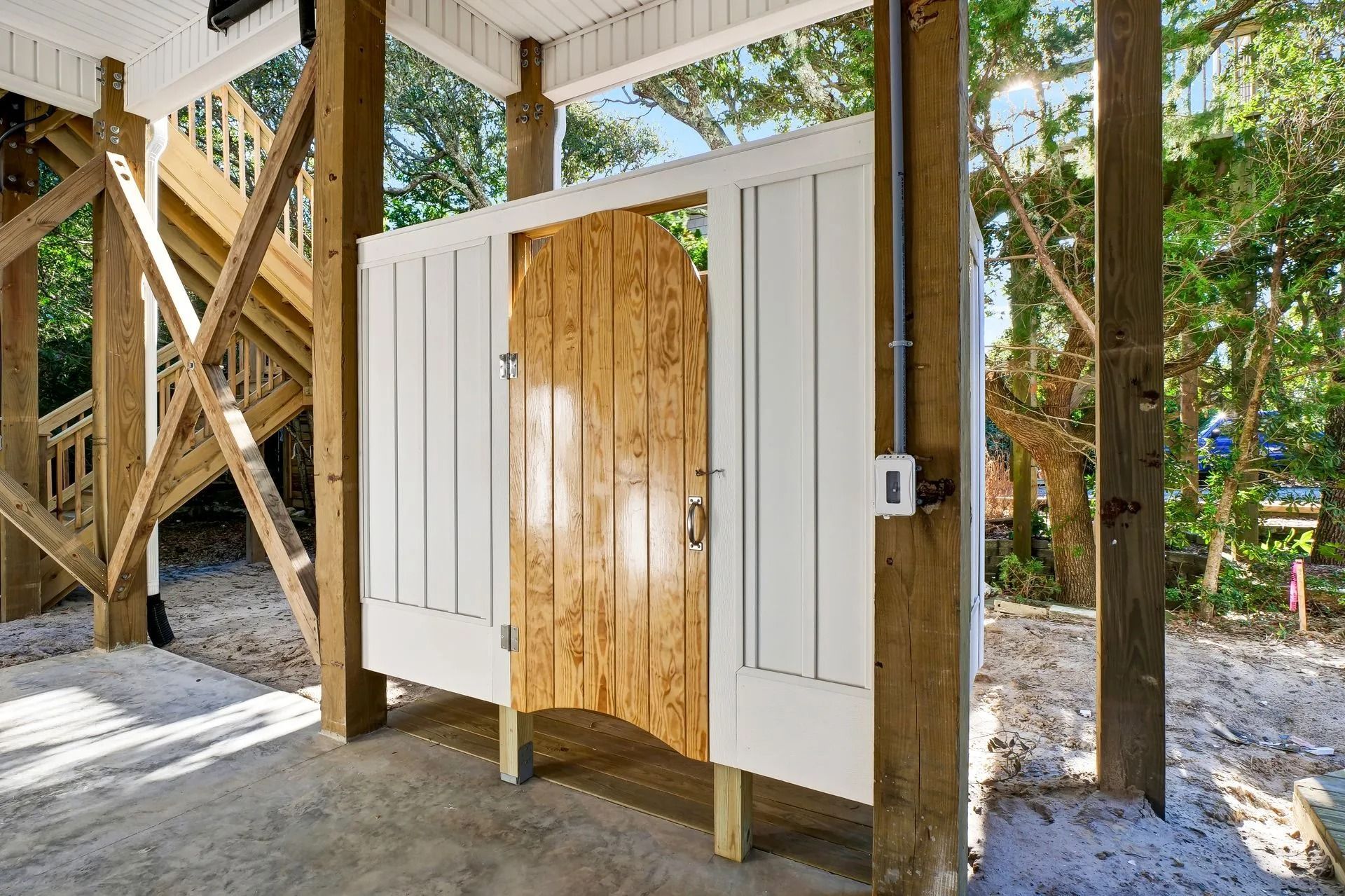 Outdoor shower under a raised wooden structure, with white siding and a wood door, on a concrete surface.