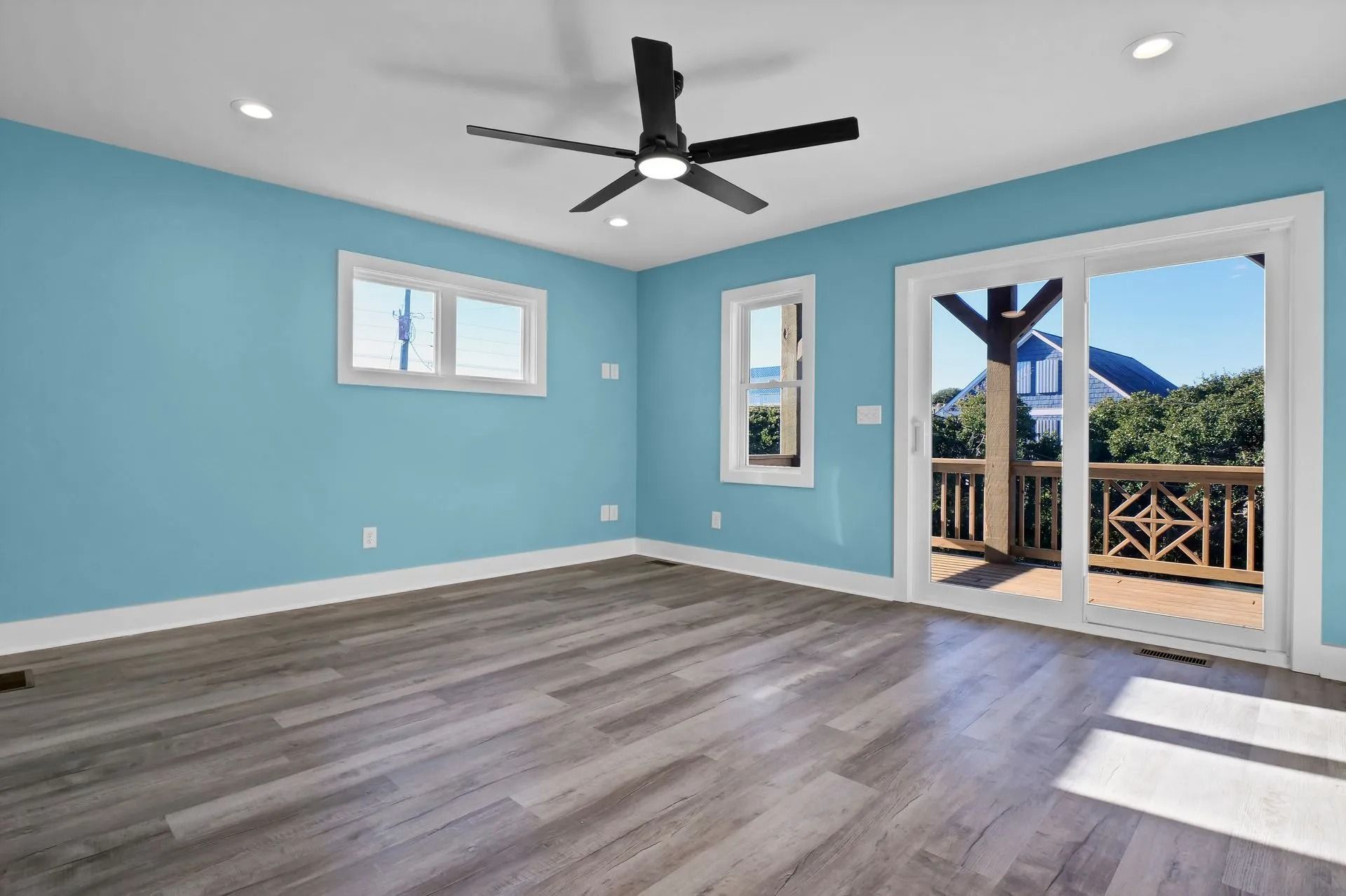 Empty room with light blue walls, gray wood-look floor, windows, sliding door to a deck, and ceiling fan.
