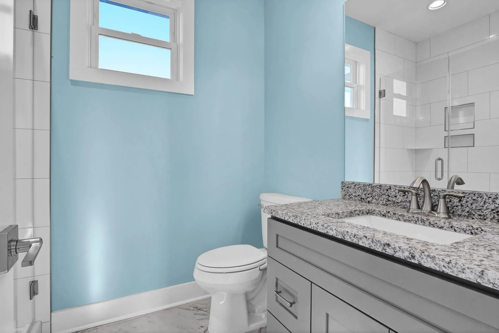 Bathroom with blue walls, white toilet, gray vanity with granite countertop, and window.
