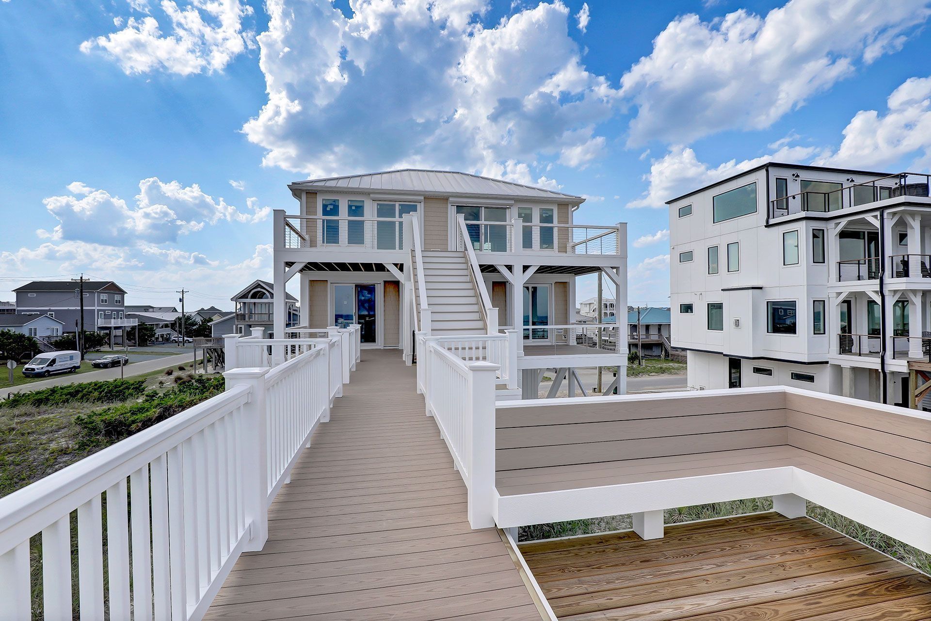 Two-story beach house with white railings, stairs, and a walkway under a blue sky with clouds.