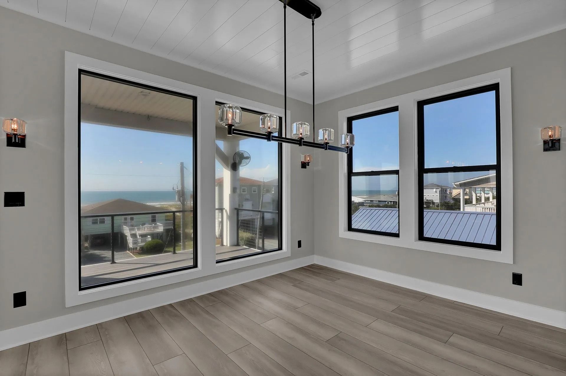 Empty room with large windows overlooking a beach and ocean, gray walls, wood floor, and chandelier.