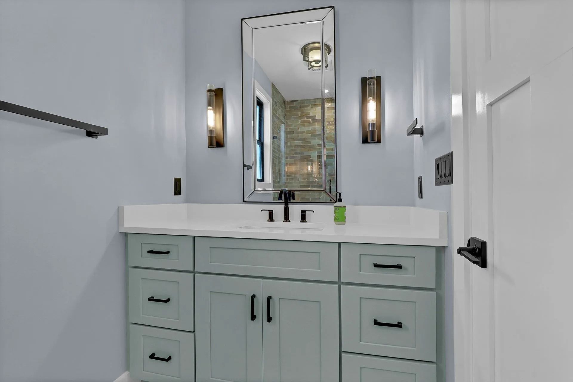 Bathroom with a light blue vanity, black fixtures, rectangular mirror, and sconce lighting.