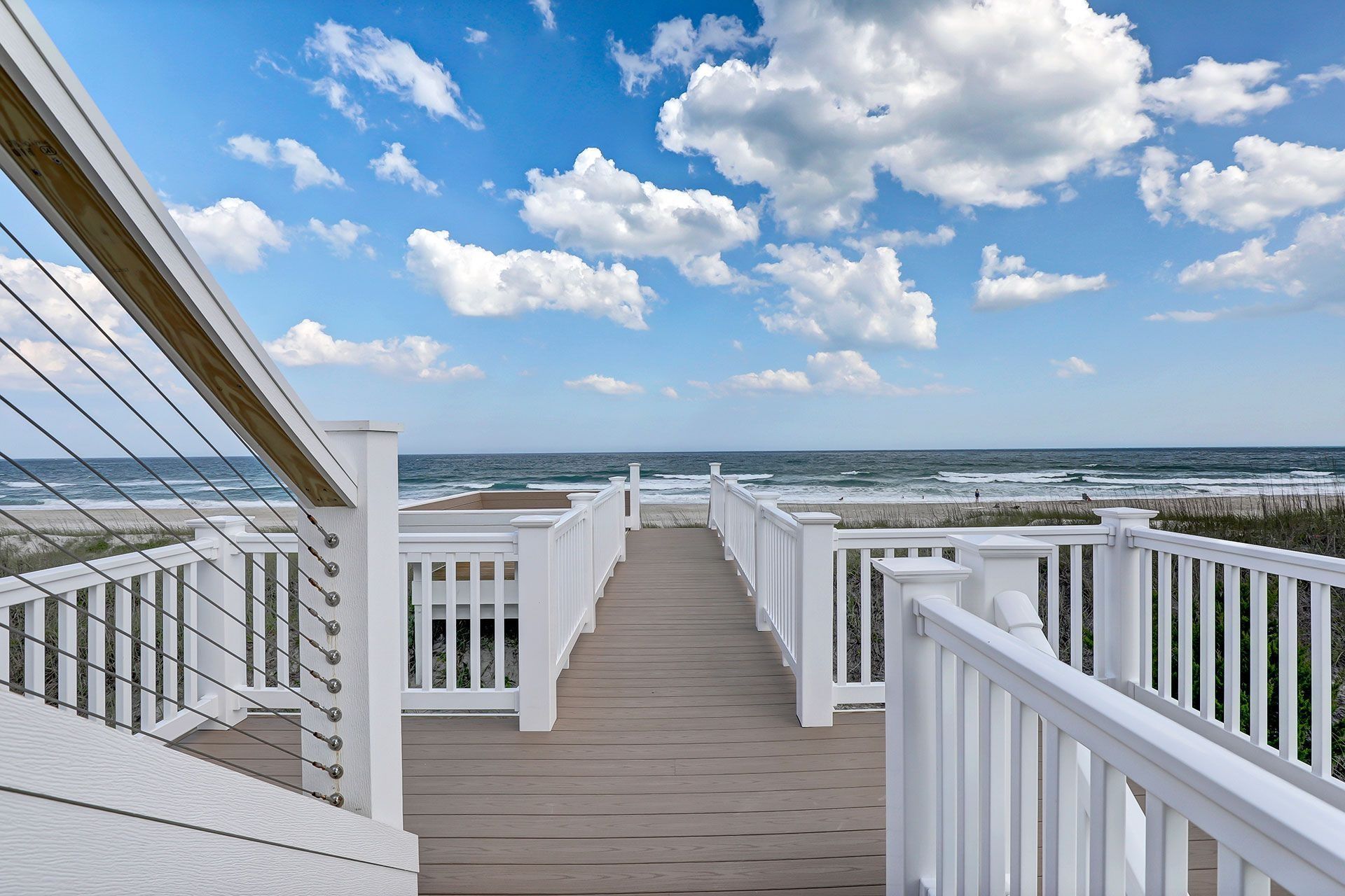 Wooden walkway with white railings leads to ocean view under a blue, cloudy sky.