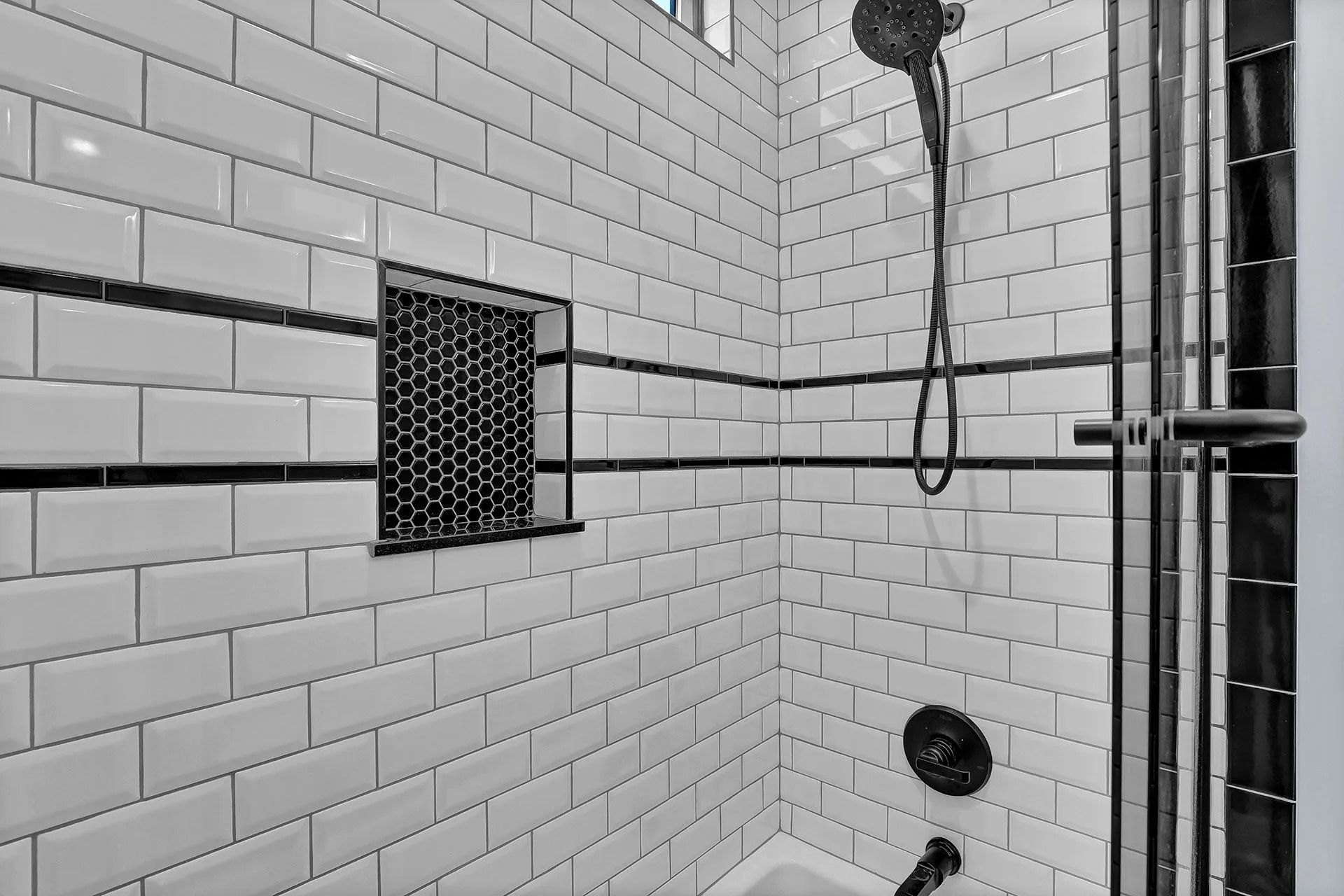 White-tiled shower with black accents. Showerhead, built-in shelf, and glass door.