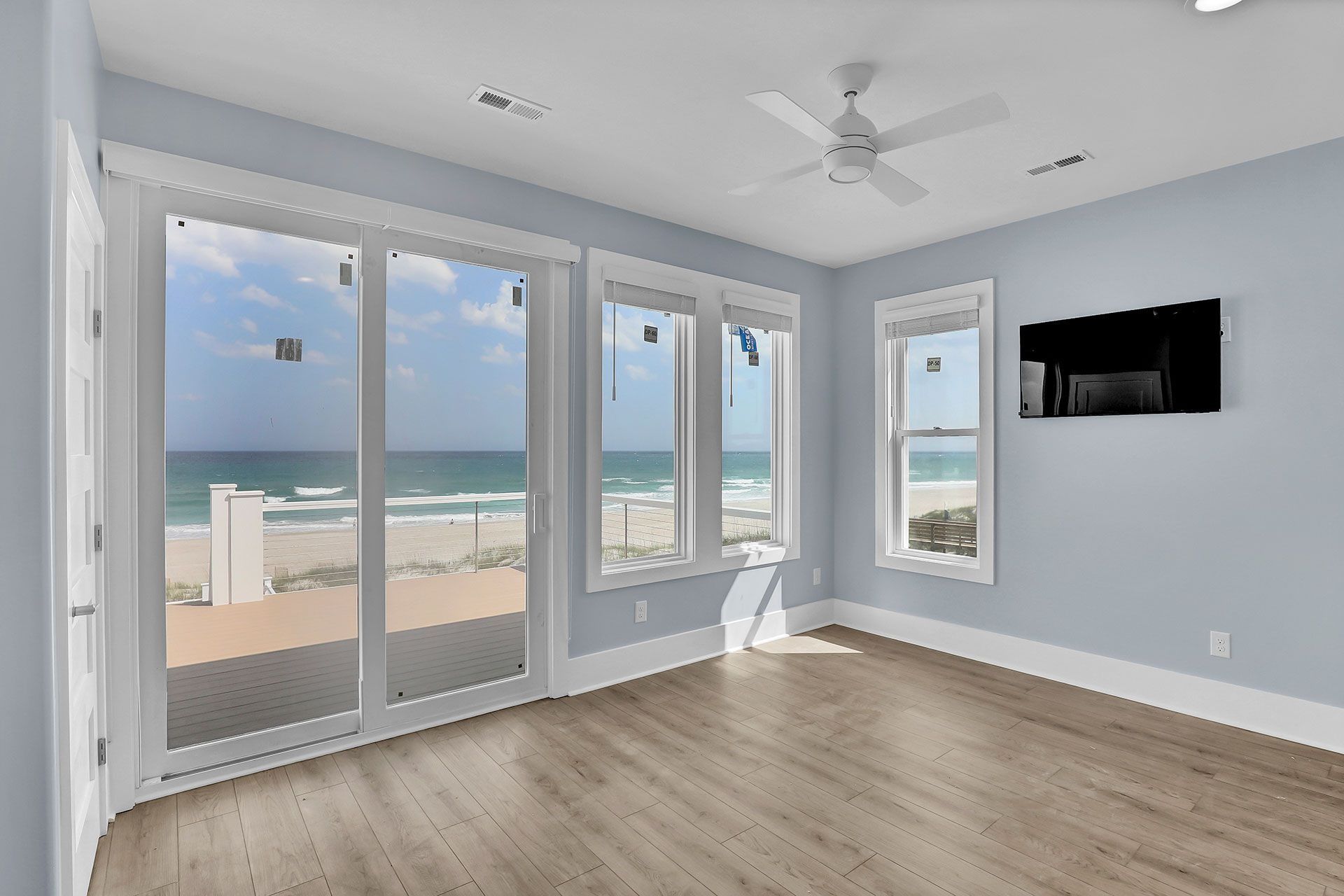 A light blue room with ocean view, large windows, wood floor, and mounted TV.
