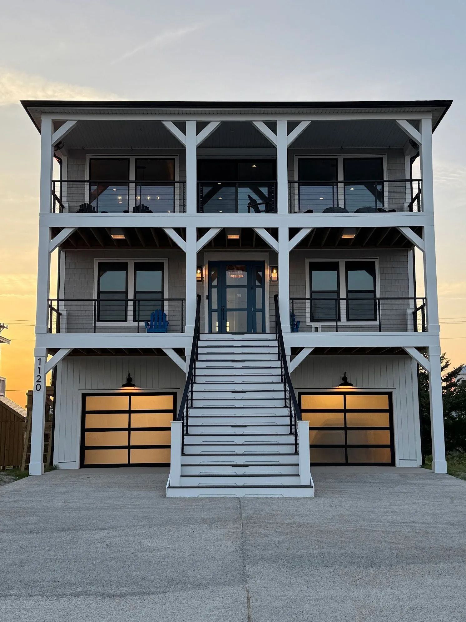 Three-story white house with balconies, black railings, and glass garage doors; stairs lead to a blue front door.