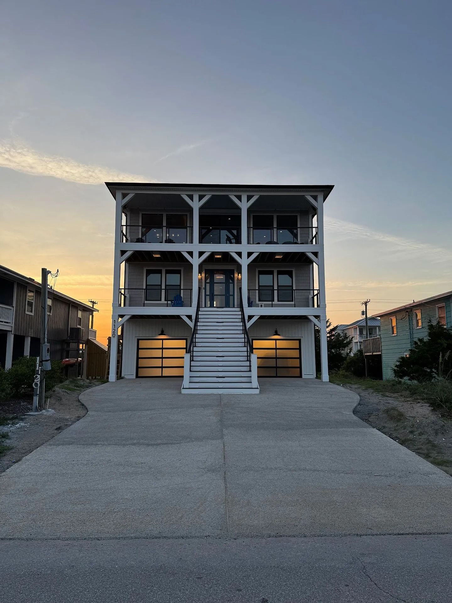 Three-story beach house with balconies, stairs, and two garage doors; gray and white with a sunset sky.