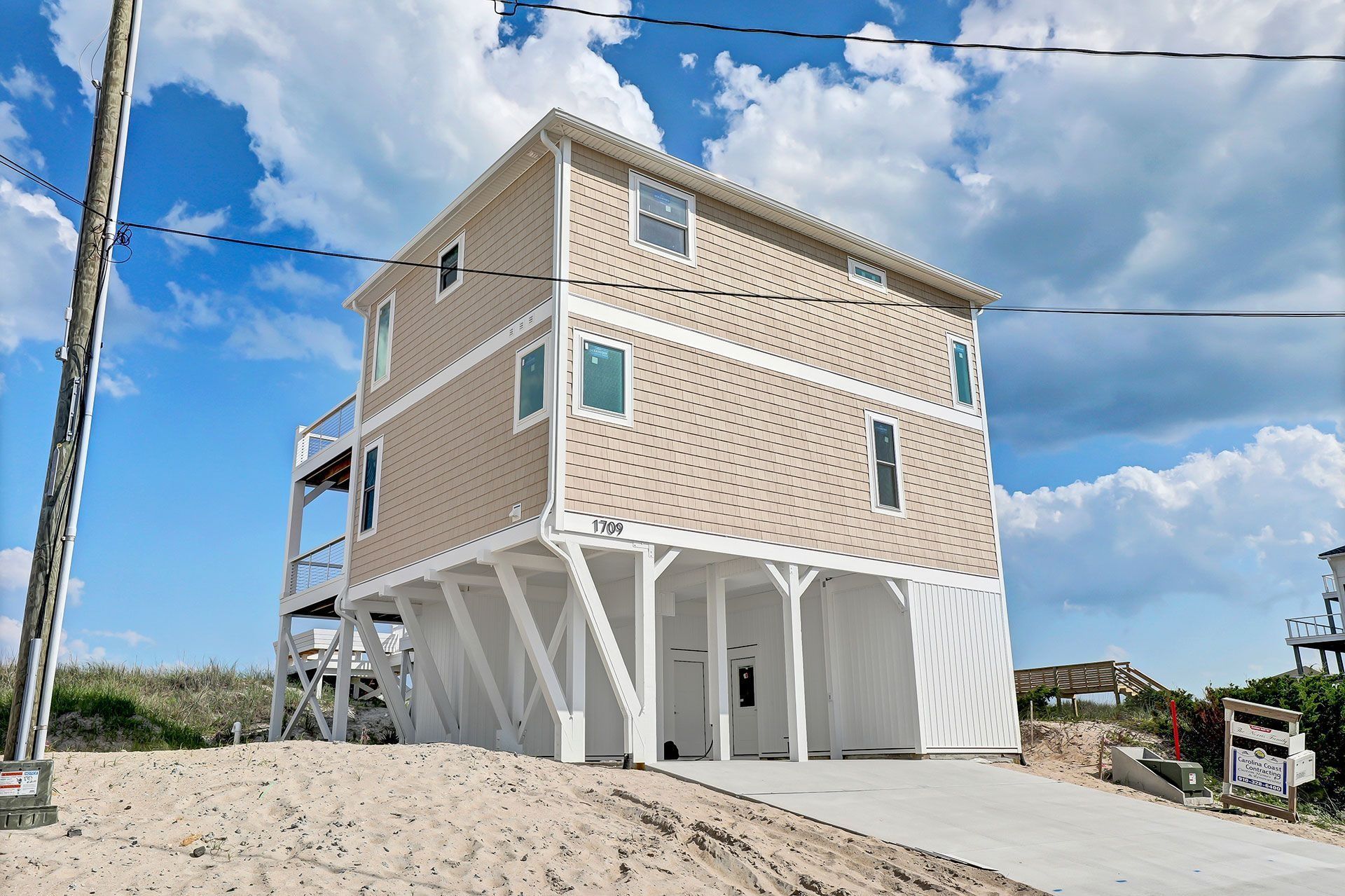 Beach house with tan siding, white supports, and a driveway, under a cloudy sky.