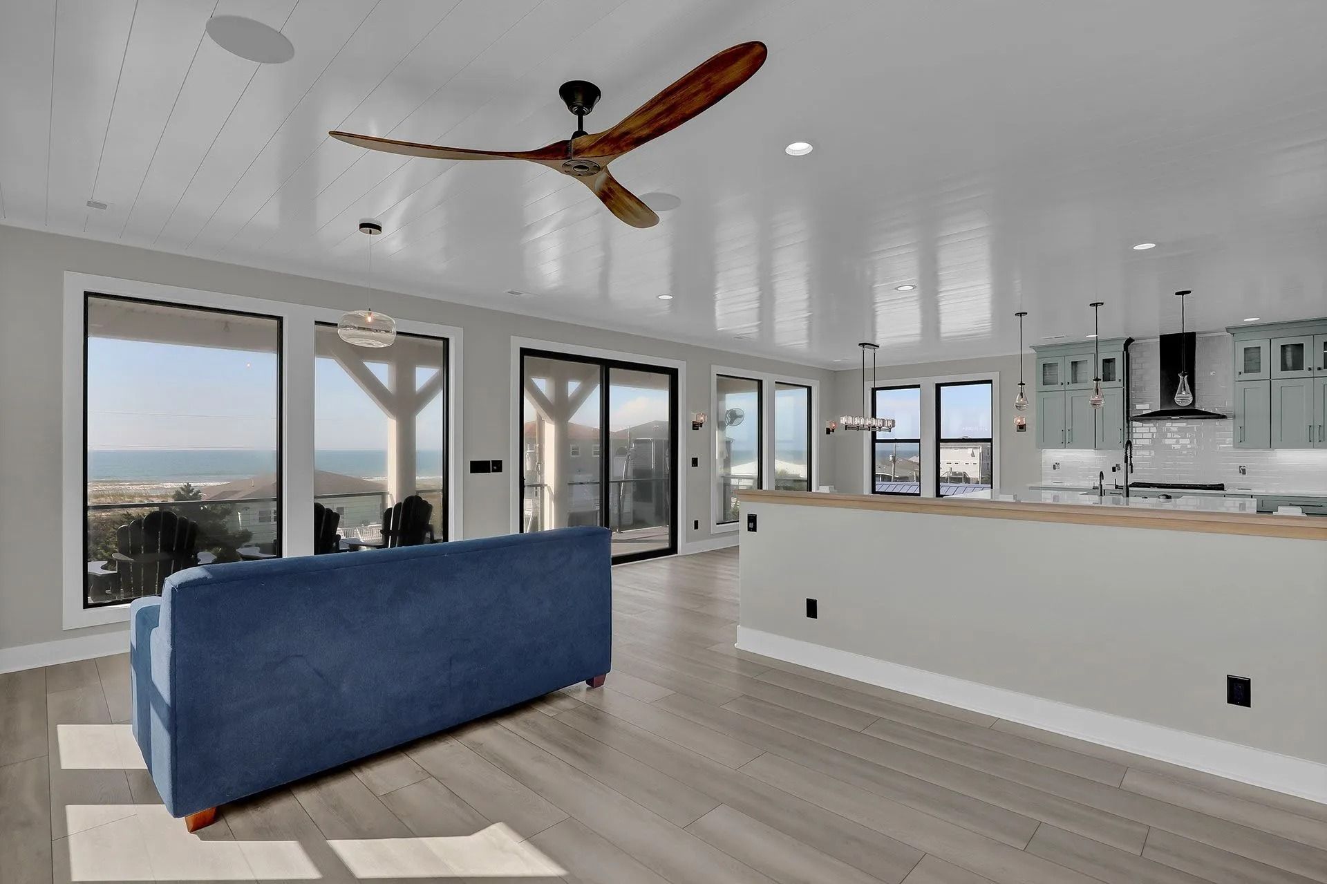 Open-concept living space with blue sofa, large windows, ceiling fan, and kitchen island. Ocean view.