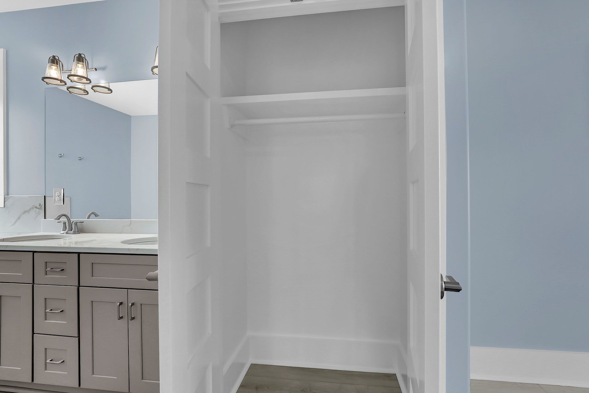 Empty white closet in a light blue bathroom. Grey vanity with double sinks in the background.