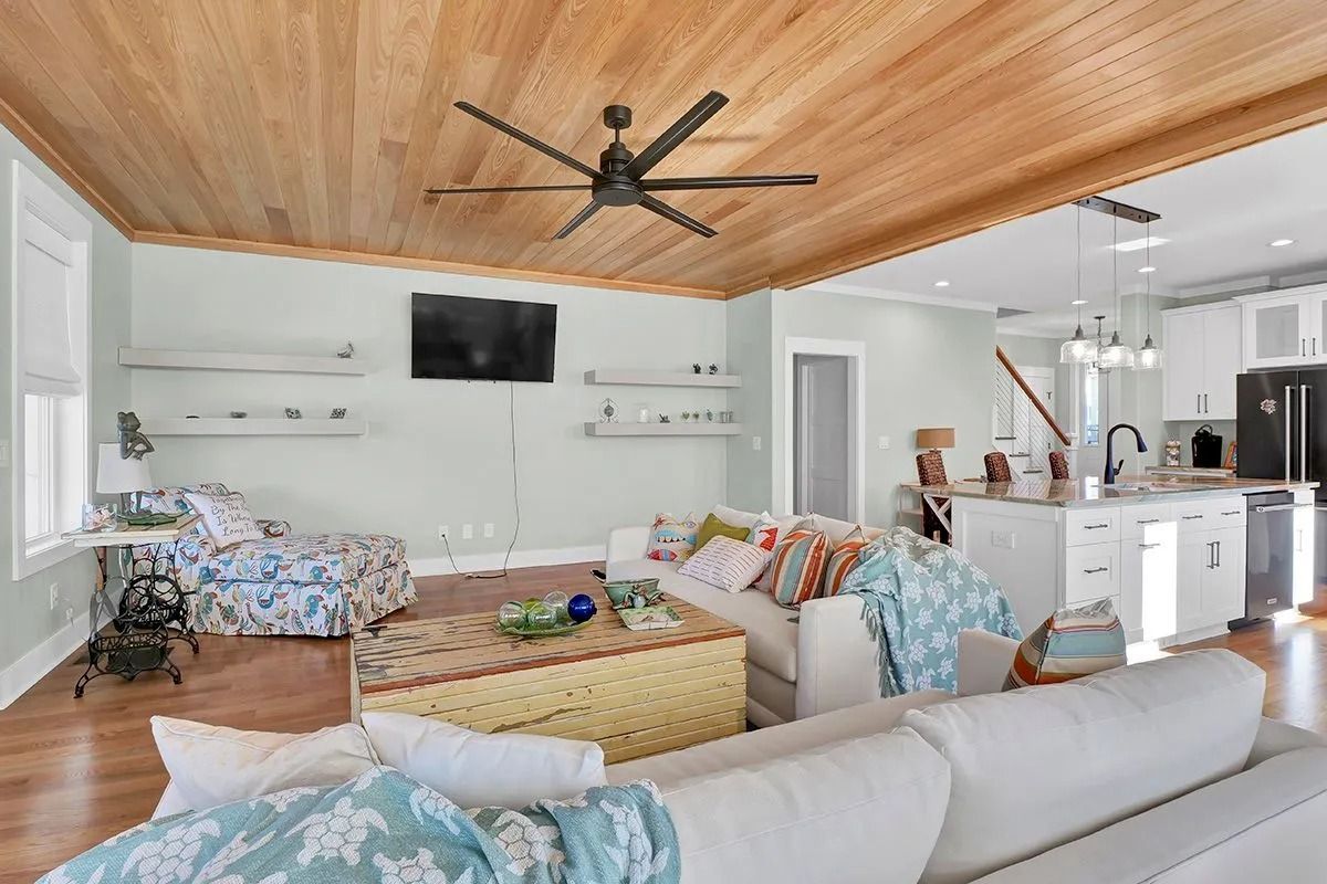 Living room with wooden ceiling, white sofa, shelves, TV, and kitchen in the background.