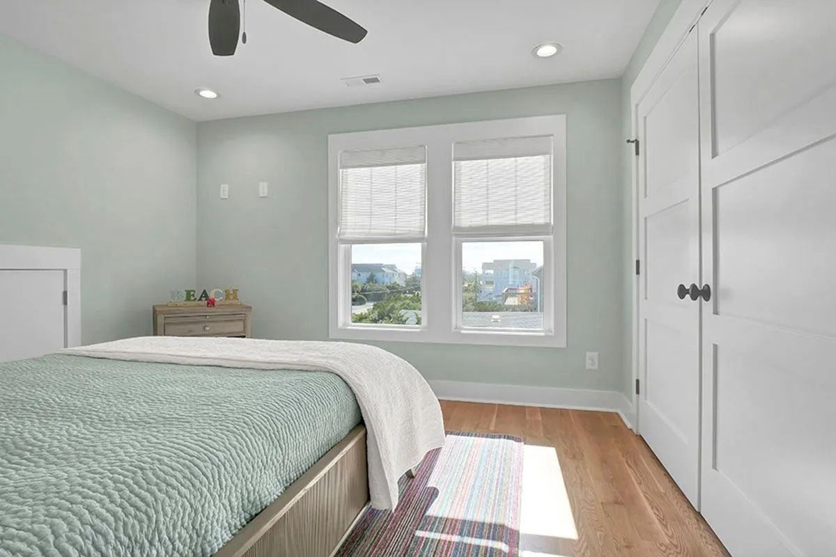 Bedroom with pale green walls, light wood floor, bed, and white window with a view.