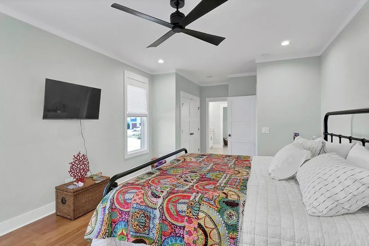 Bedroom with bed, TV, ceiling fan, and window. Colorful bedspread and white walls.