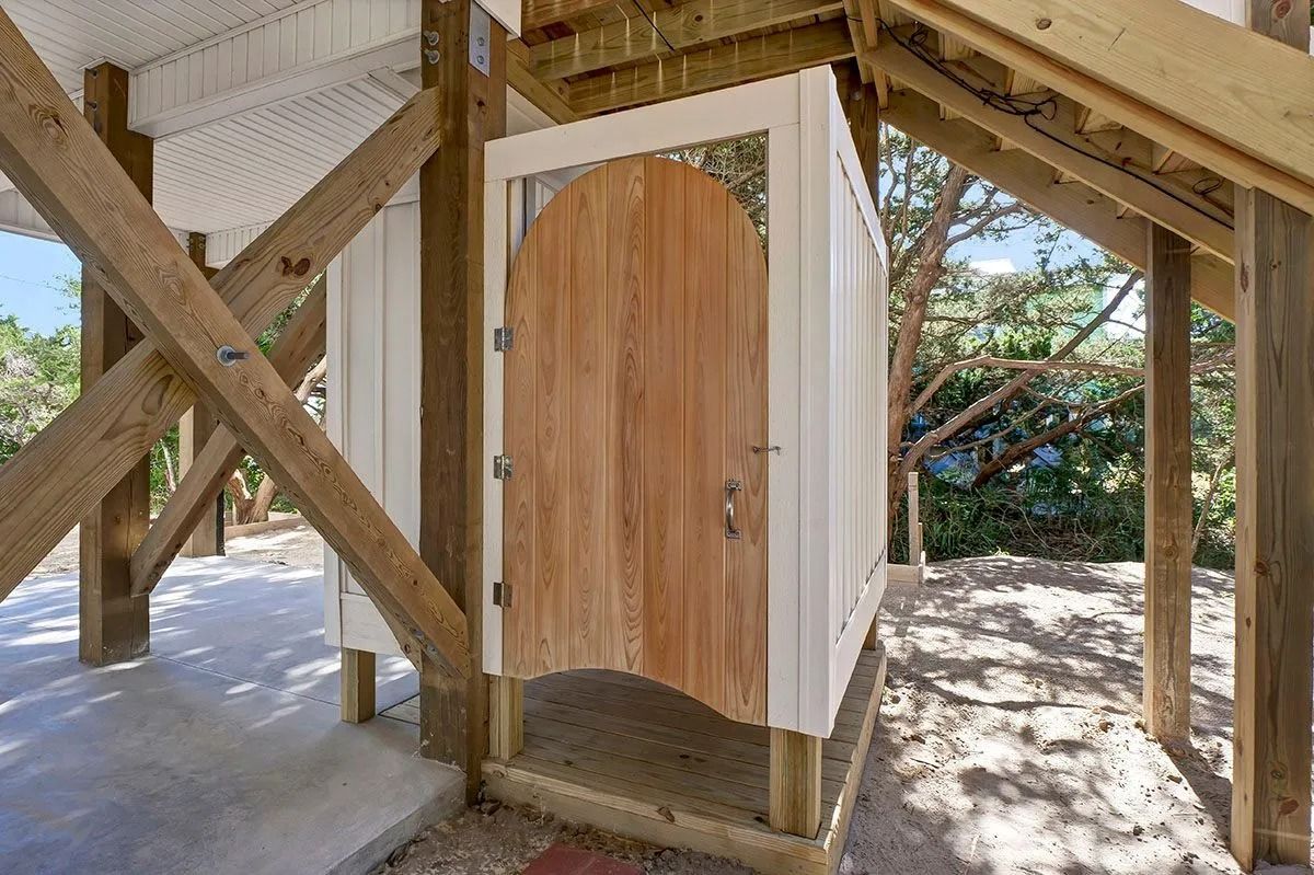 Outdoor wooden shower under a raised structure; light wood door, white walls, sunny setting.