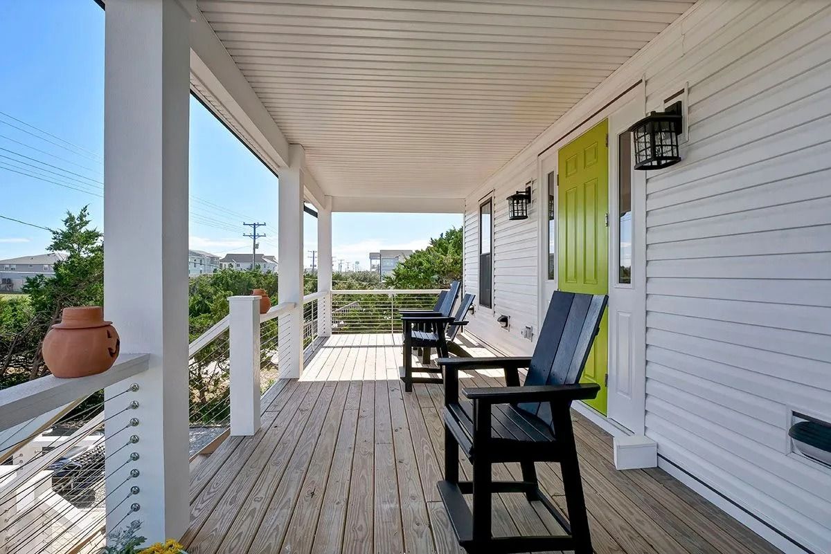 Porch of a white beach house with black chairs, a green door, and view of the neighborhood.
