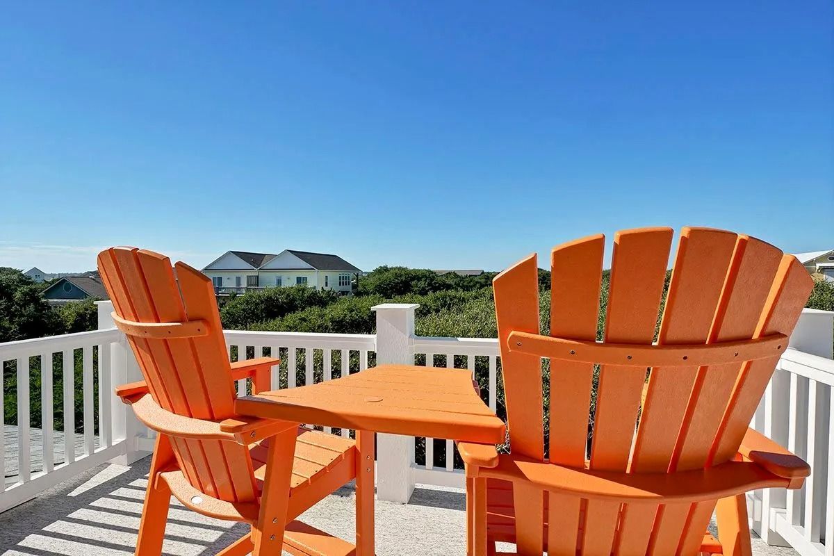 Two orange Adirondack chairs on a balcony with a small table, overlooking a house and blue sky.