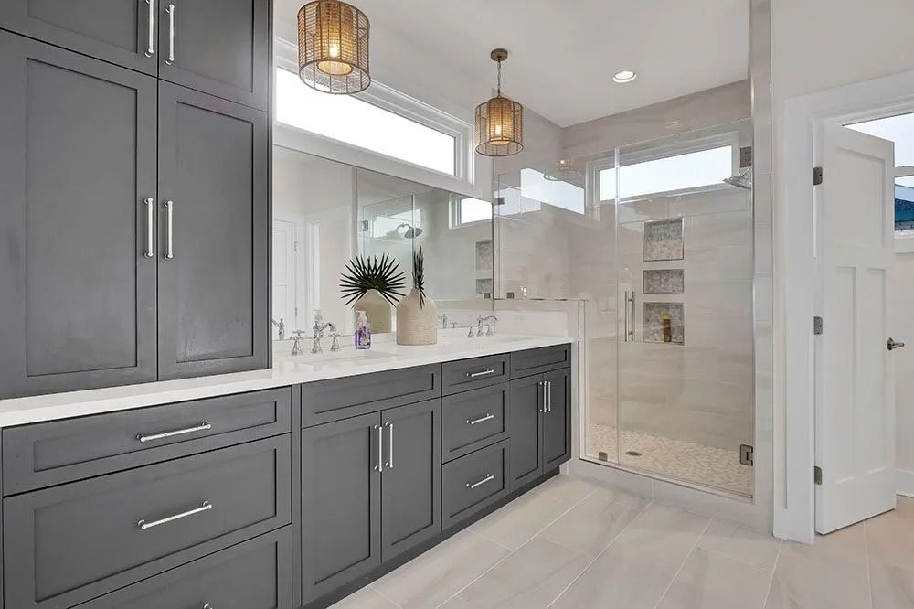 Gray and white bathroom with large vanity, tall storage, and glass shower. Two pendant lights hang.