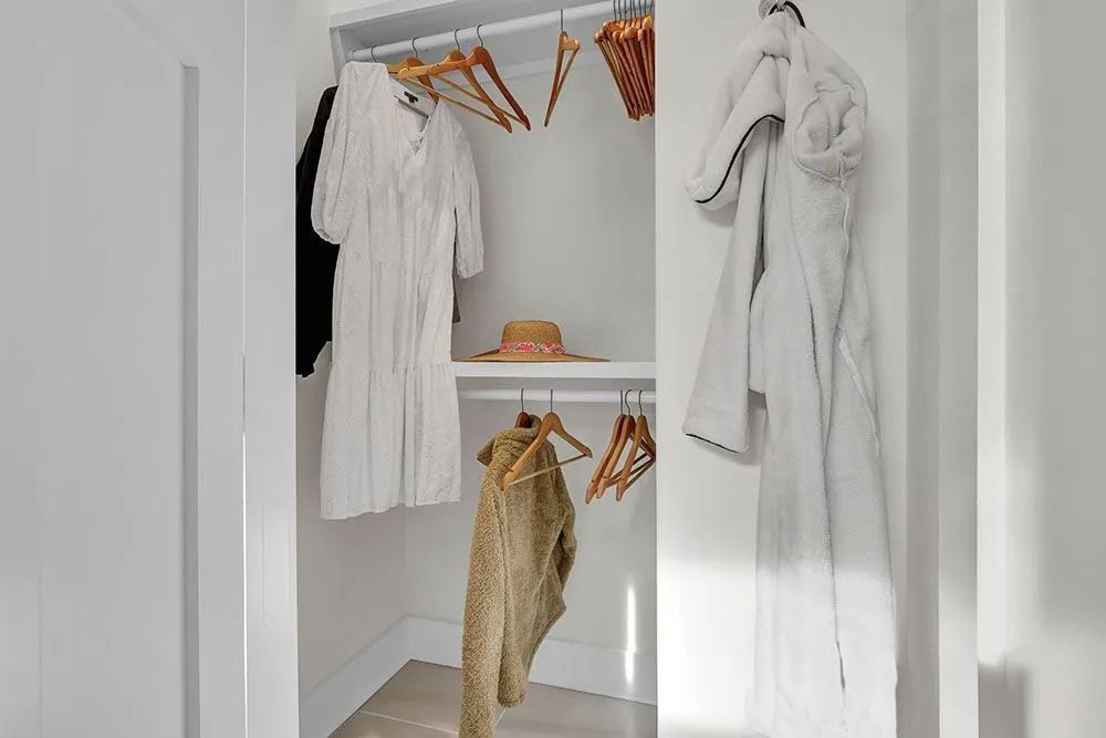 A small white closet with clothes hanging. A straw hat sits on a shelf.