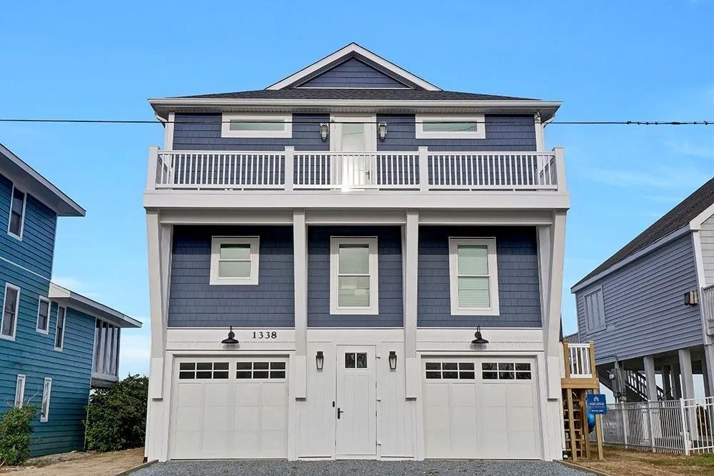 Two-story blue beach house with a balcony, garage, and white trim against a blue sky.