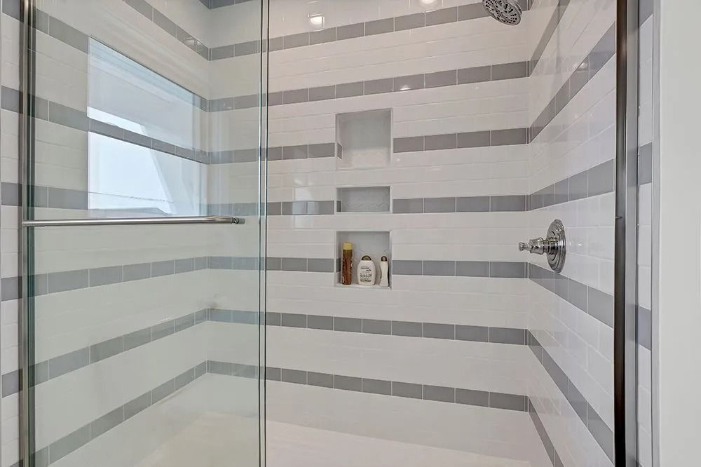 Shower with white and gray striped tile, glass doors, and recessed shelves holding toiletries.