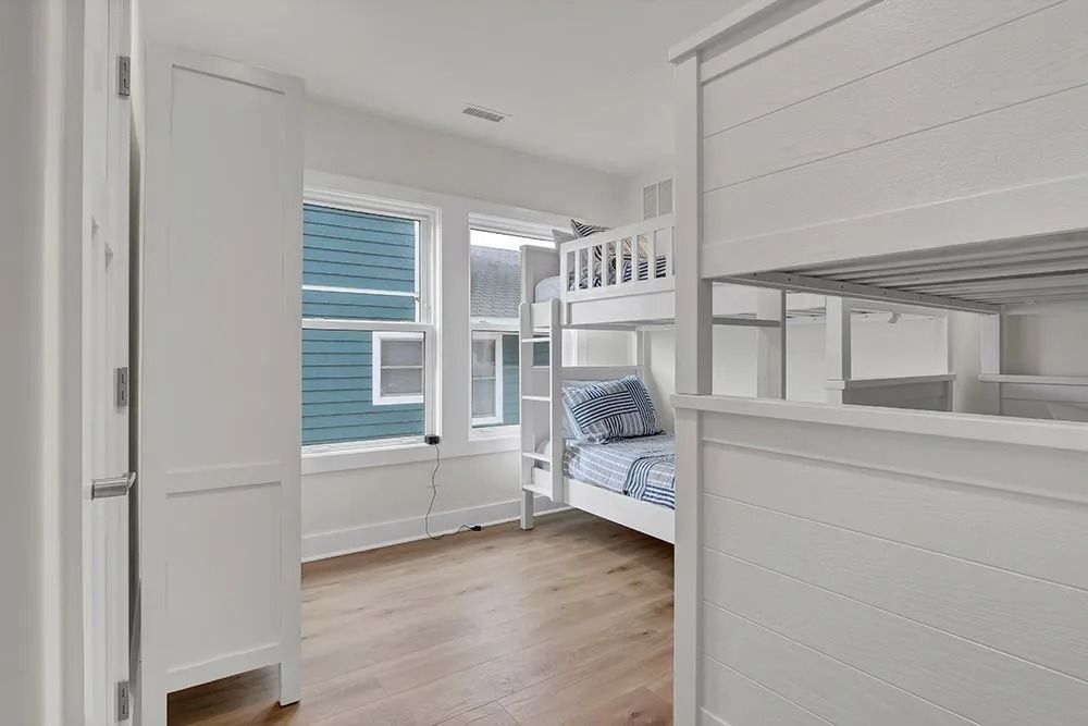 White bunk beds in a bright room with light wooden floors, a white wardrobe, and two windows.
