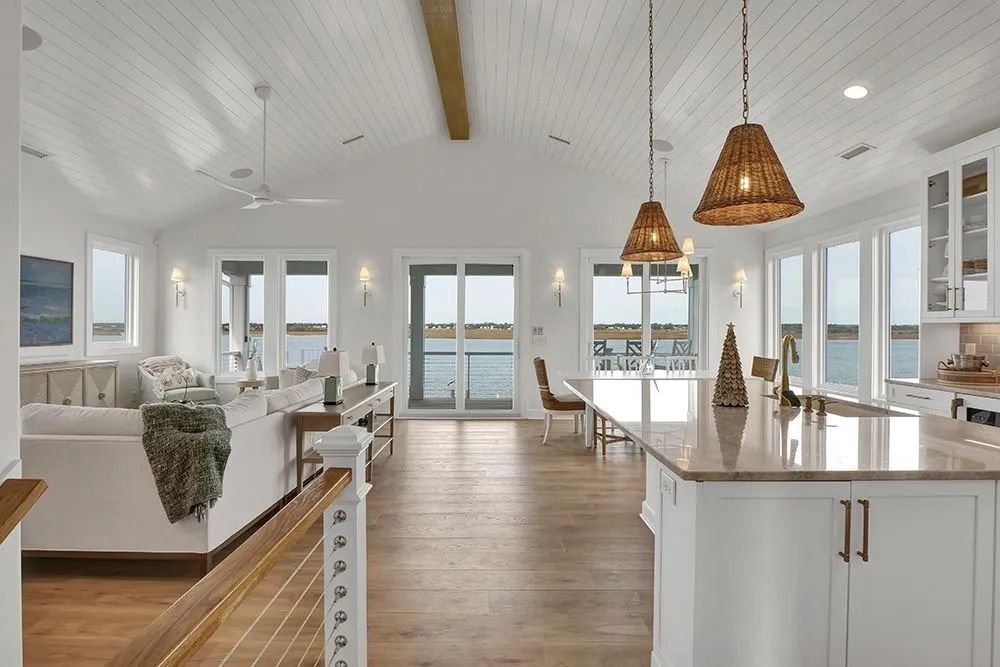Open-concept kitchen and living area with water views. White walls, wooden floors, pendant lights.