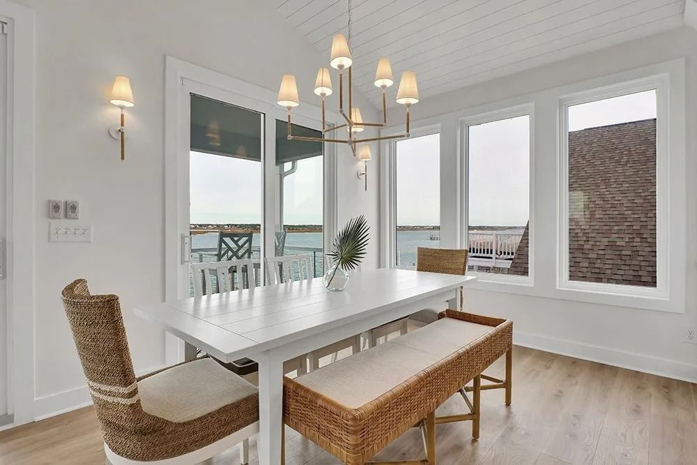 Dining room with white table, bench, and chairs, large windows overlooking water.