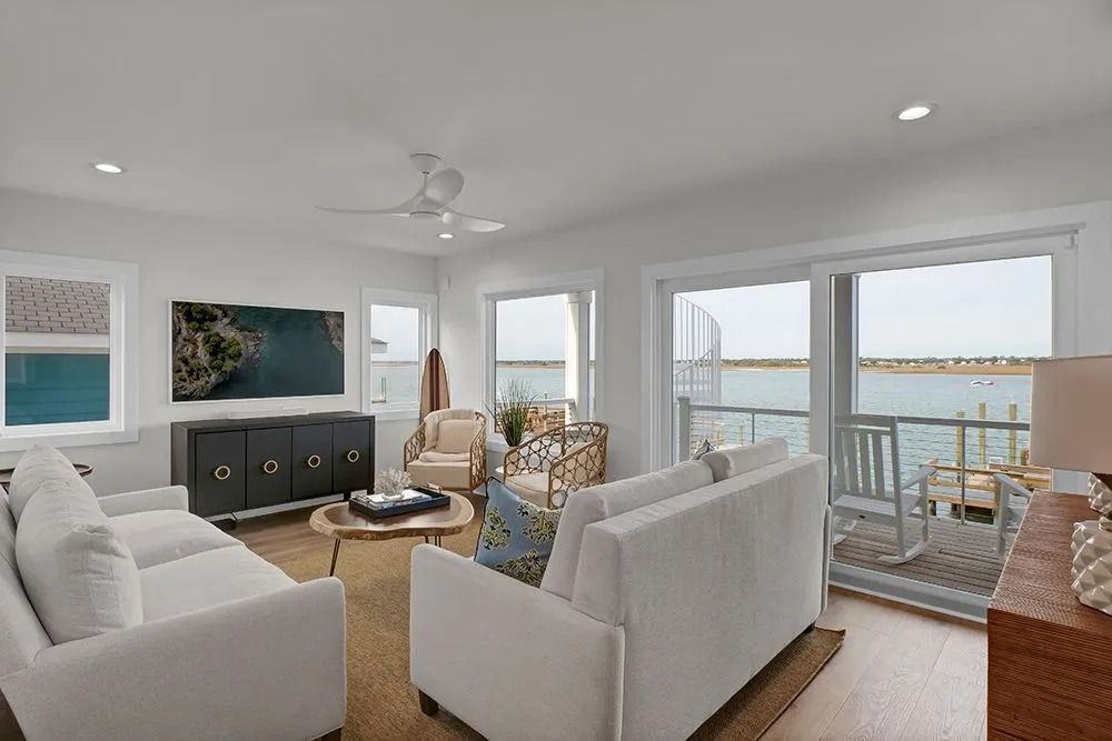Bright living room with white sofas, water view from sliding glass doors, and light wood furniture.