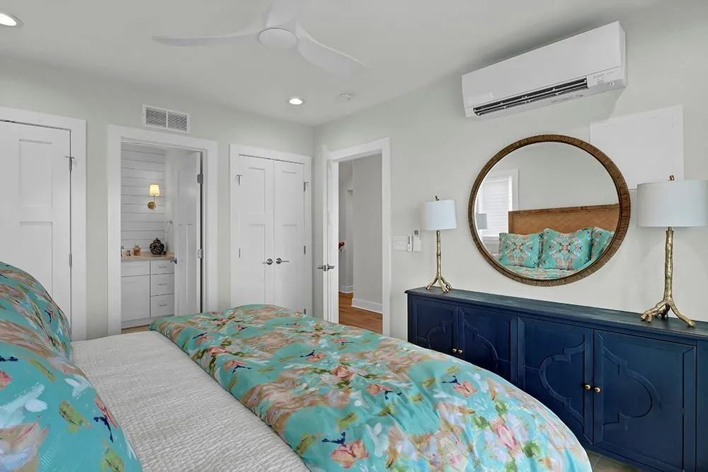 Bedroom with blue floral bedspread, navy dresser, round mirror, and air conditioner.