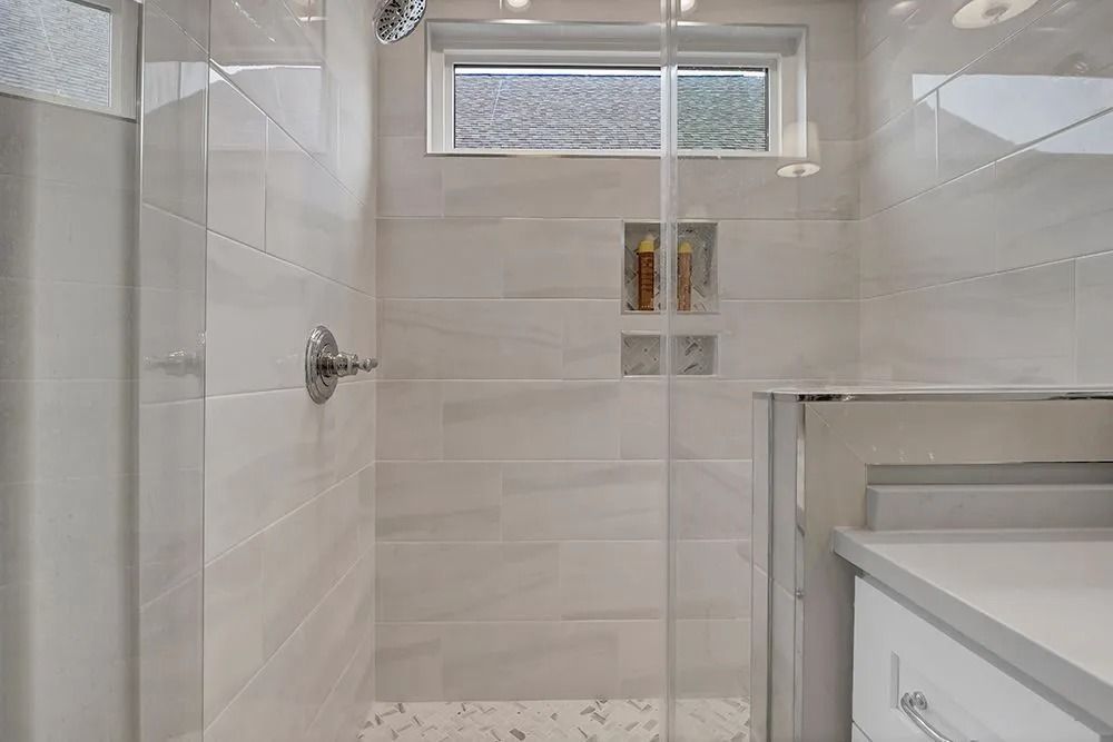 Modern shower with white tiled walls, glass door, window, built-in shelves, and chrome fixtures.