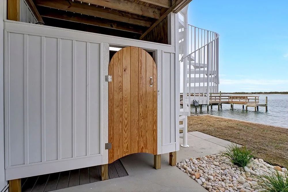 Outdoor shower with wooden door, white siding, next to a spiral staircase and dock on the water.
