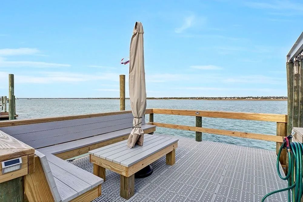 Dockside seating area with a table, bench, and umbrella, overlooking calm water and sky.
