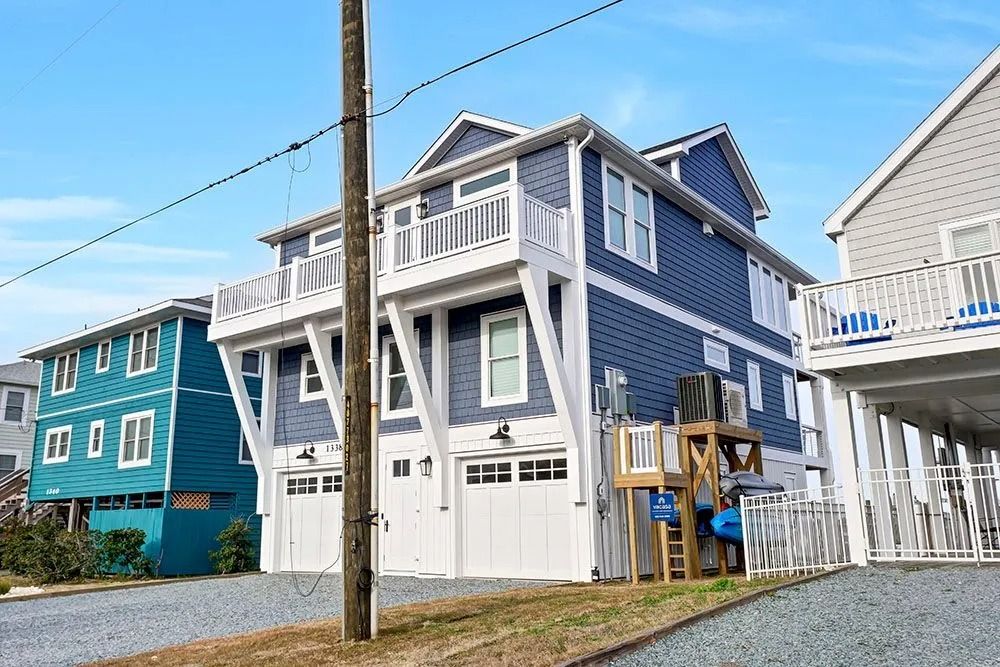 Two-story blue house on stilts with white trim and balconies. Garage doors at the base.