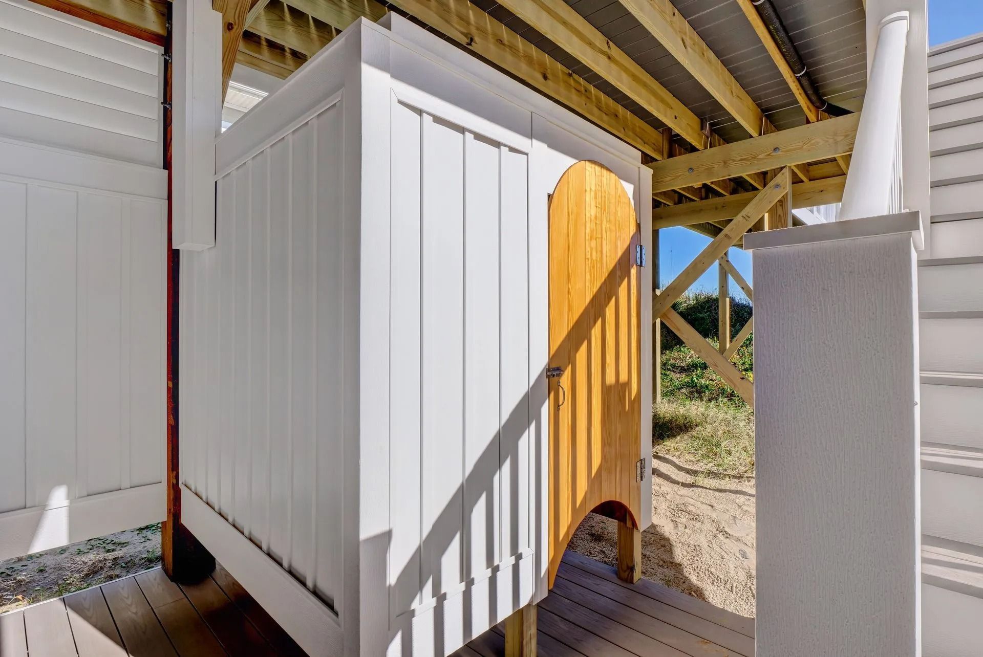 Outdoor shower with white siding and a wooden door beneath a deck.