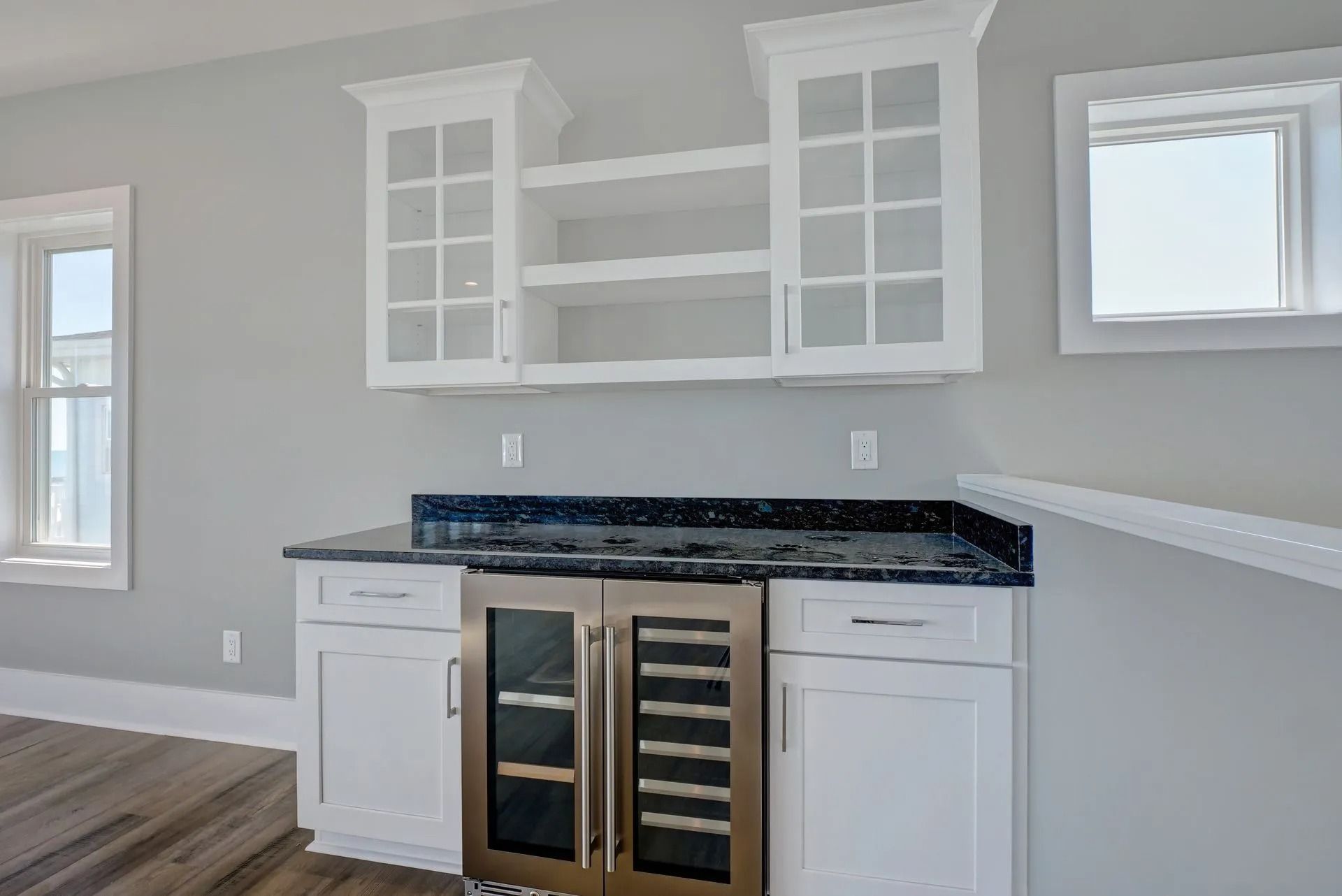 White cabinets, wine fridge, and open shelving beneath a light gray wall, with windows.