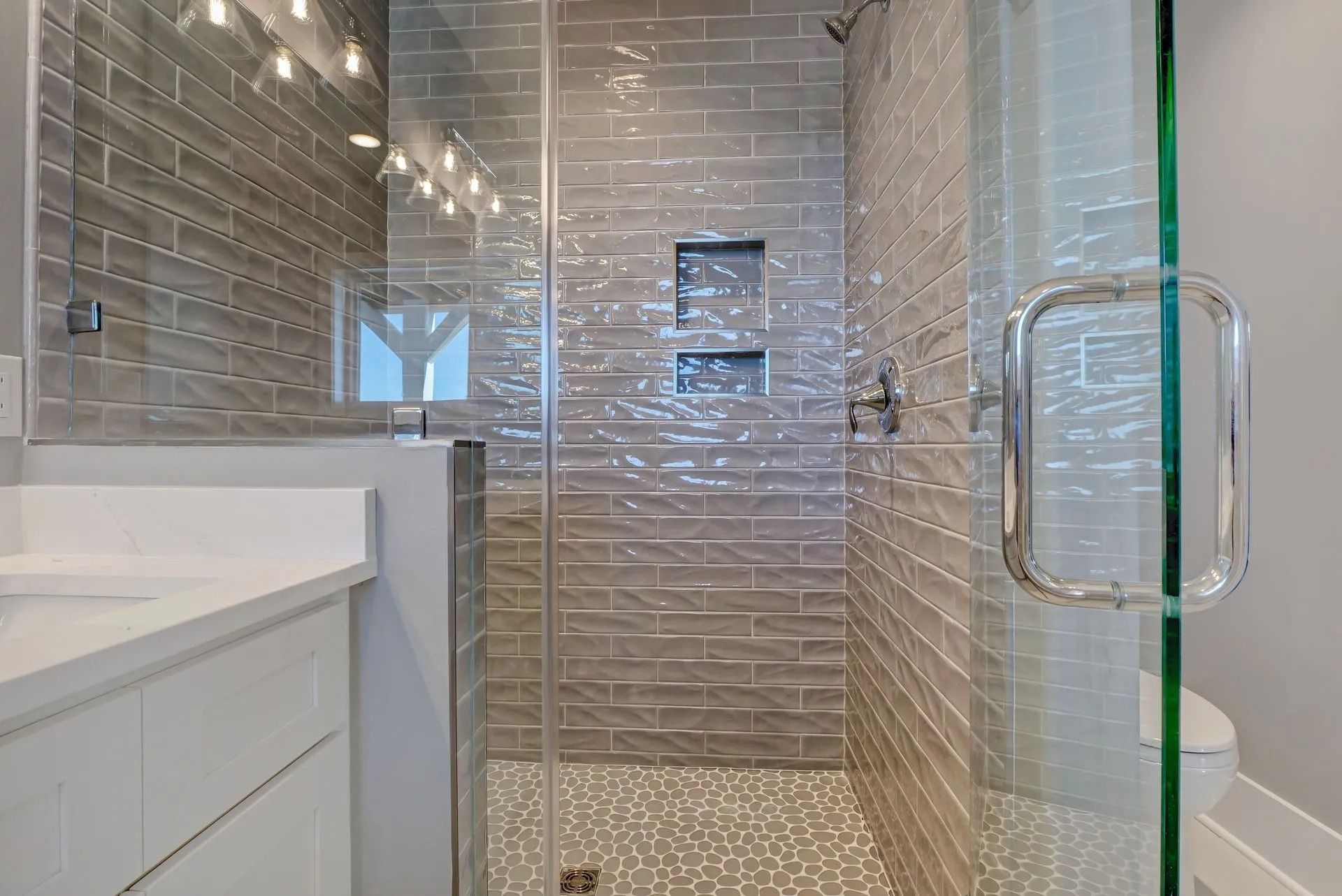 Gray-tiled shower with glass door, pebble floor, and white vanity in a modern bathroom setting.
