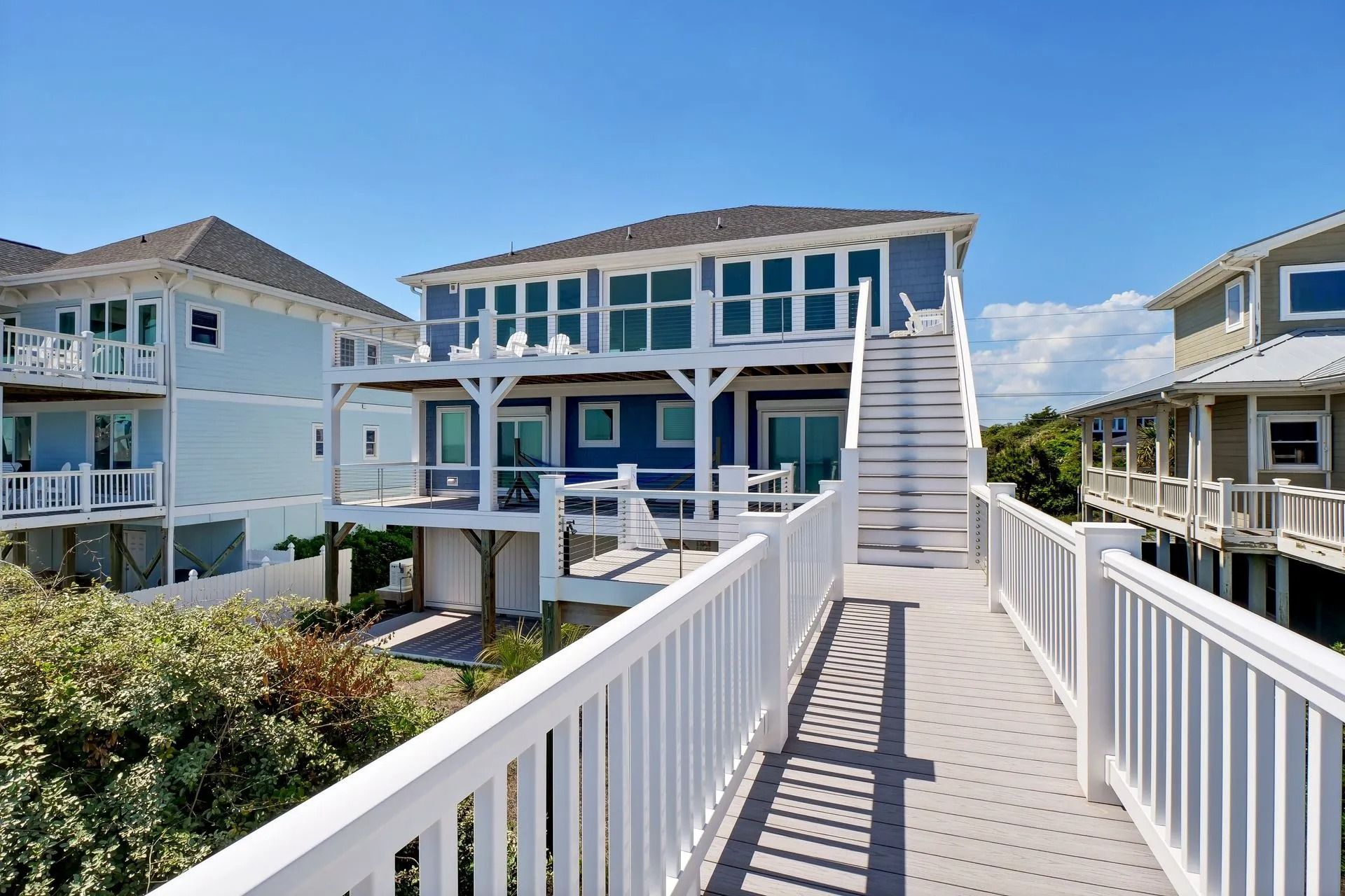 Beach house with blue siding, connected to a walkway with white railing, under a clear blue sky.