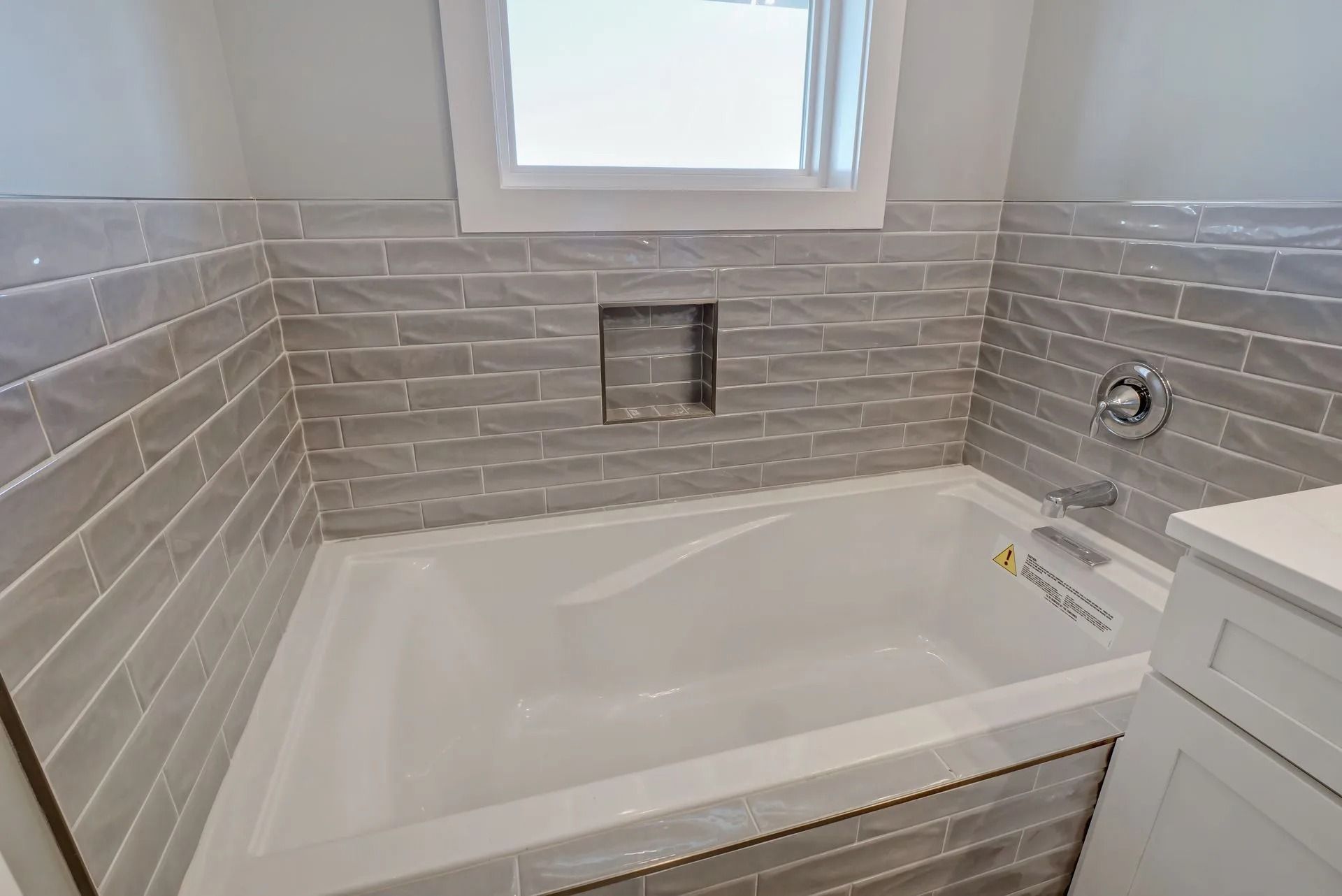 Gray tiled bathroom with white bathtub, niche, and window.