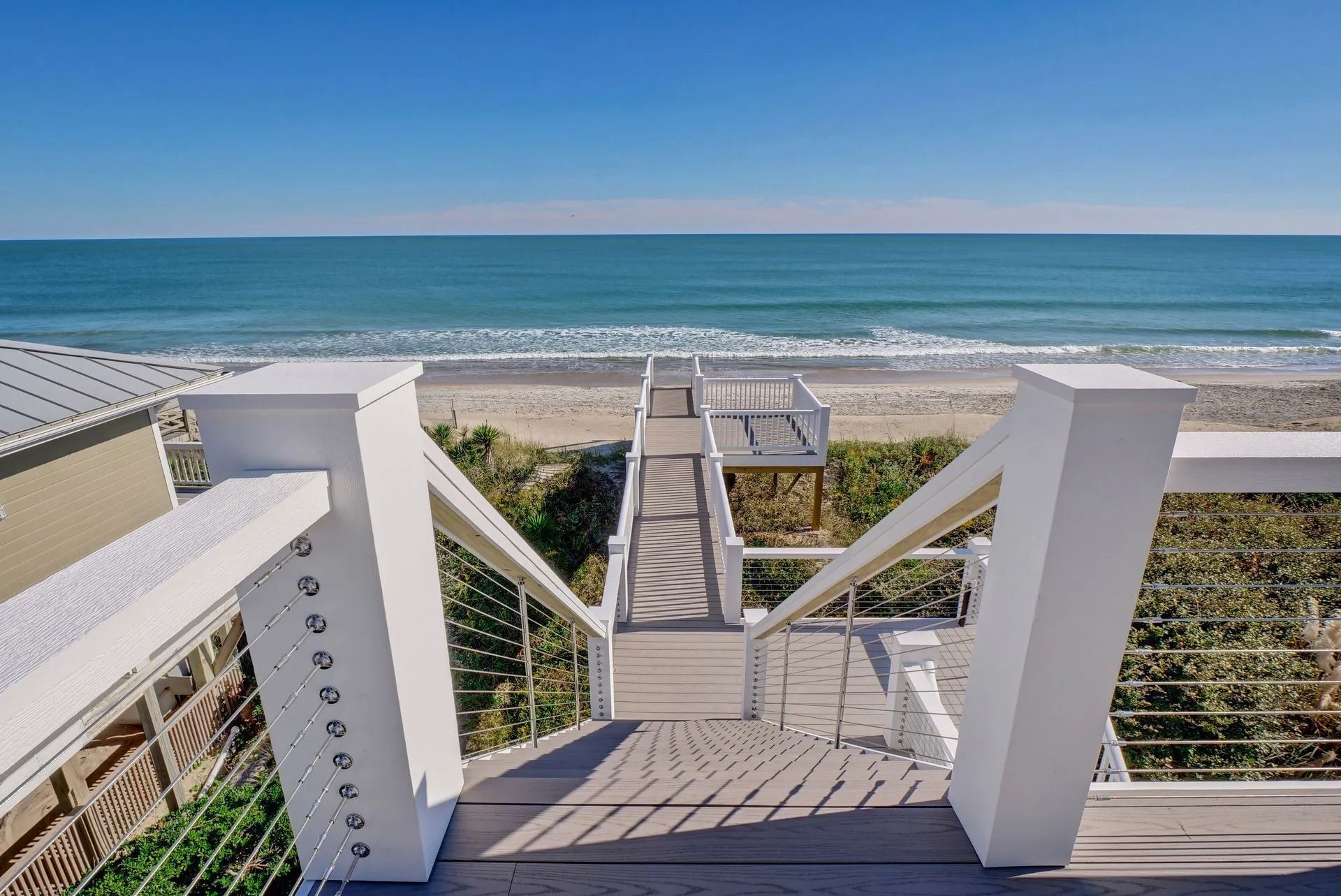 Staircase leading to a beach and ocean view. White railing, gray deck, blue sky and water.
