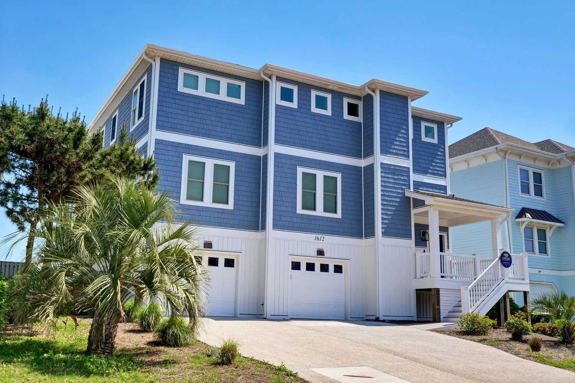 Blue multi-story beach house with white trim, driveway, and garage doors under a clear blue sky.
